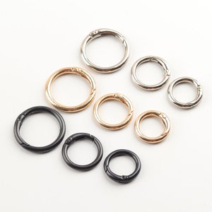 May include: A collection of round spring snap rings in various sizes and finishes. The rings are in gold, silver, and black. The clasps are open, showing the spring mechanism. These are commonly used for keychains, jewelry, and crafting projects.