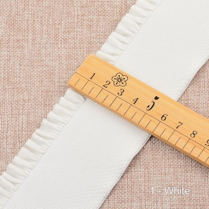 May include: White cotton trim with a ruffled edge. The trim is measured with a wooden ruler, showing it is 5 inches wide. The text "1 - White" is visible below the ruler.