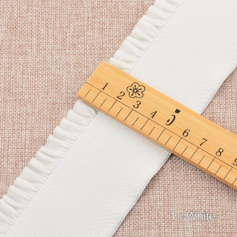 May include: White cotton trim with a ruffled edge. The trim is measured with a wooden ruler, showing it is 5 inches wide. The text "1 - White" is visible below the ruler.