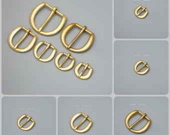 Puede incluir: Una colección de hebillas de cinturón en forma de D, de color latón, en varios tamaños. Las hebillas varían de pequeñas a grandes, con dimensiones internas de 14 mm, 17 mm, 20 mm, 25 mm y 32 mm. Fondo gris.