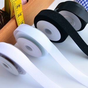 May include: Spools of white and black hook and loop tape, also known as Velcro, are displayed. The white tape is unrolled, revealing its textured surface. A yellow measuring tape is visible in the background, suggesting a sewing or crafting theme.