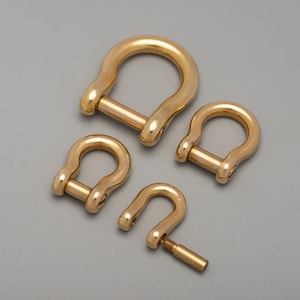 May include: Four gold-colored D-shackles of varying sizes are arranged on a gray background. One shackle is partially disassembled, showing the removable pin. The shackles have a rounded, U-shaped design with a pin closure.