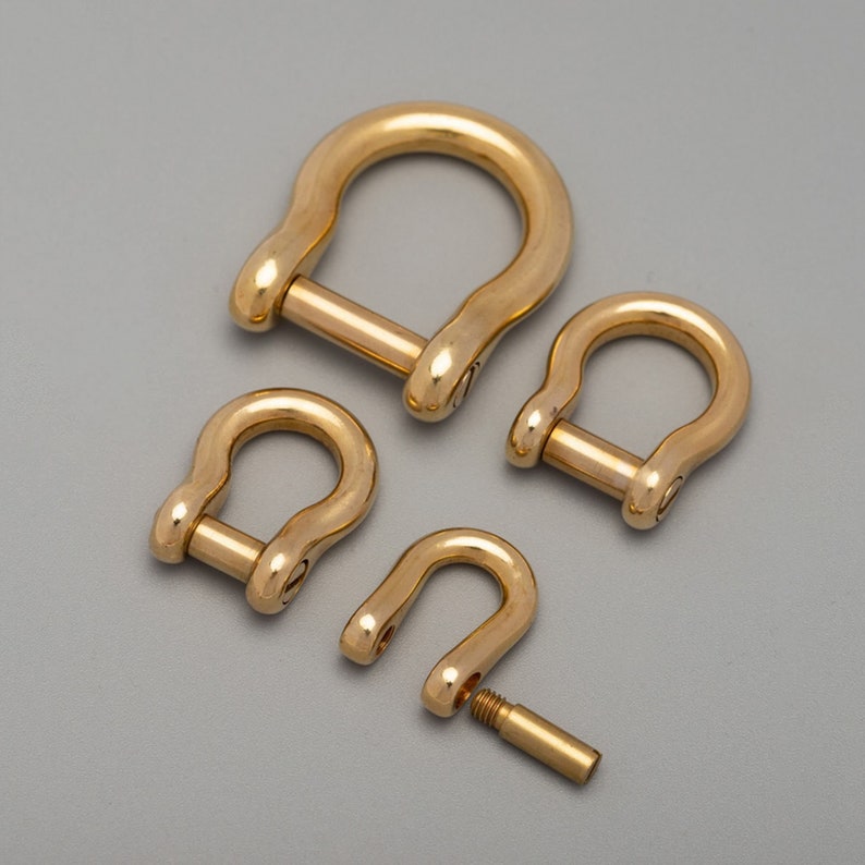 May include: Four gold-colored D-shackles of varying sizes are arranged on a gray background. One shackle is partially disassembled, showing the removable pin. The shackles have a rounded, U-shaped design with a pin closure.
