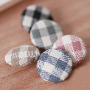 Puede incluir: Una colección de botones cubiertos de tela en varios colores, incluyendo blanco y negro, gris y blanco, marrón y blanco, azul y blanco, y rosa y blanco. Los botones tienen un patrón de cuadros clásico.