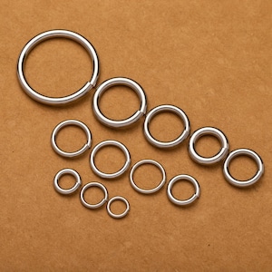 May include: A set of 11 silver jump rings in various sizes, arranged in a descending order from largest to smallest on a brown surface.