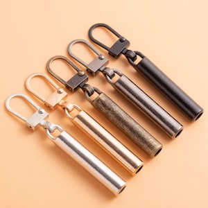 May include: Five zipper pulls in different finishes: silver, gold, antique brass, gunmetal, and black. Each pull has a loop at the top and a cylindrical body.