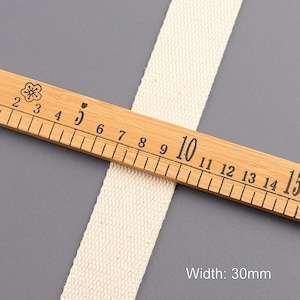 May include: A close-up of a natural white cotton webbing measuring 30mm wide. The webbing is laid across a wooden ruler with black markings.
