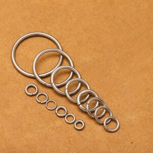 May include: A set of silver metal jump rings of various sizes, arranged in a descending order of size on a brown surface.