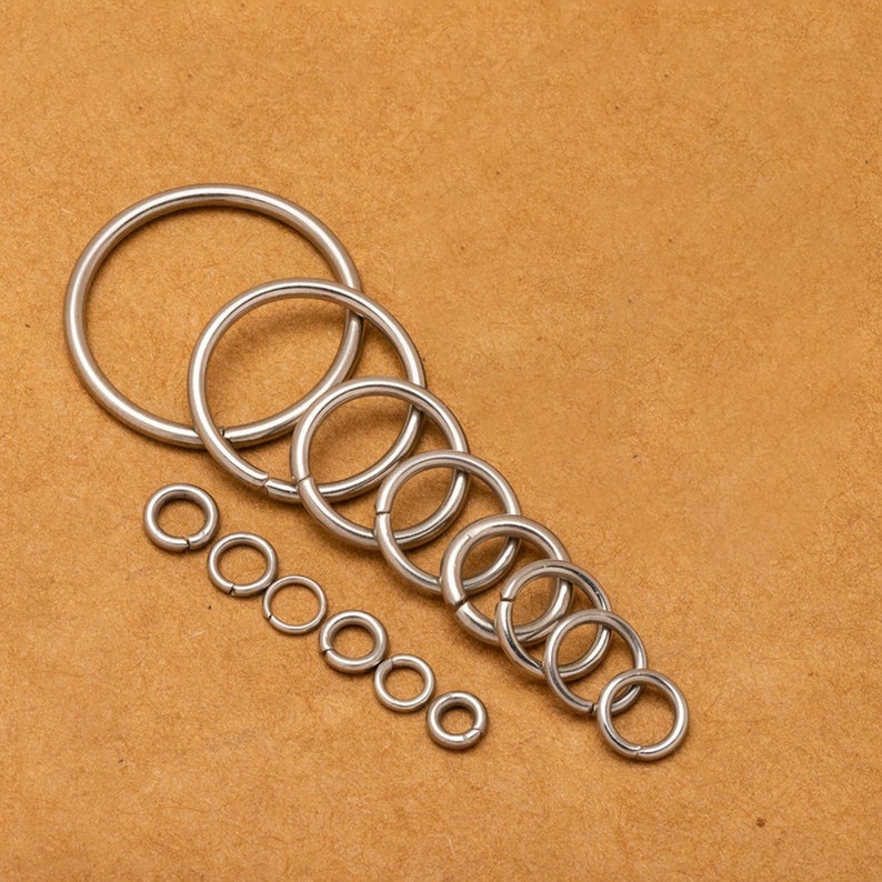 May include: A set of silver metal jump rings of various sizes, arranged in a descending order of size on a brown surface.