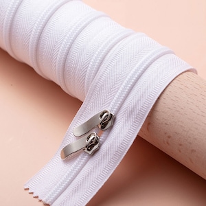May include: A roll of white zipper tape with two silver zipper pulls. The zipper is tightly wound, showing the teeth and the fabric tape. The background is a light pink color.