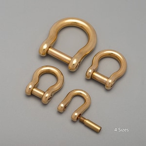 May include: Four brass D-shackles of varying sizes are arranged on a gray surface. The shackles are U-shaped with a removable pin. The text "4 Sizes" is in the lower right corner. The metal has a polished, golden finish.