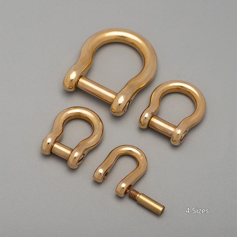 May include: Four brass D-shackles of varying sizes are arranged on a gray surface. The shackles are U-shaped with a removable pin. The text "4 Sizes" is in the lower right corner. The metal has a polished, golden finish.