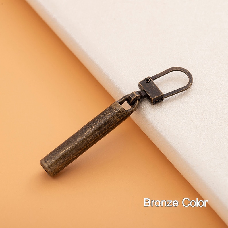 May include: A bronze-colored zipper pull with a cylindrical shape and a small loop at the end.