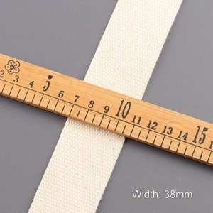 May include: A close-up of a natural white cotton webbing measuring 38mm wide. The webbing is laid on a wooden ruler with black markings.