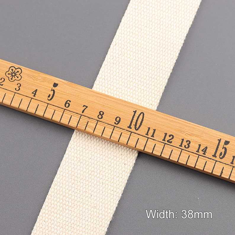 May include: A close-up of a natural white cotton webbing measuring 38mm wide. The webbing is laid on a wooden ruler with black markings.