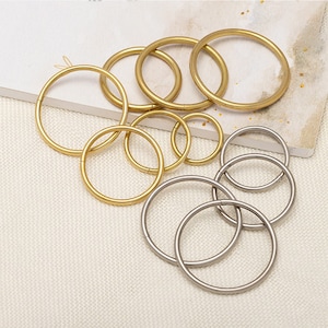 May include: Assortment of gold and silver metal rings of varying sizes. The rings are arranged in a random pattern on a beige fabric surface.