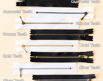 May include: A close-up of six different zipper pulls with varying metal finishes. The zipper pulls are arranged in pairs, with each pair featuring a black and white zipper tape. The metal finishes include gunmetal, bronze, gold, and silver.