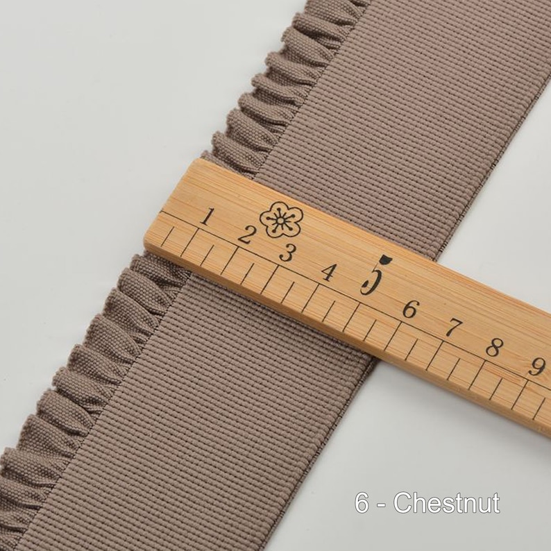 May include: A close-up of a brown ruffled trim ribbon with a wooden ruler measuring 5 inches. The ribbon is labeled "6 - Chestnut".