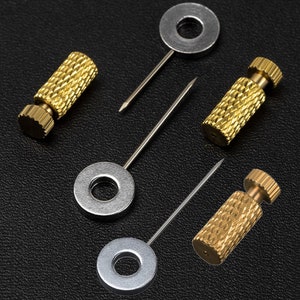 Könnte beinhalten: Vier silberne Metallringe mit einem kleinen Loch in der Mitte, die jeweils an einer dünnen silbernen Nadel befestigt sind. Die Nadeln sind an goldfarbenen zylindrischen Sockeln mit einer strukturierten Oberfläche befestigt.