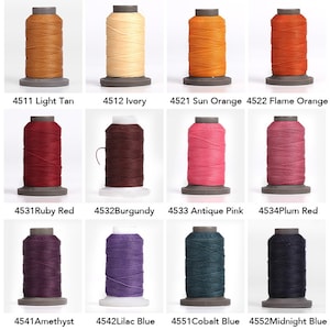 Puede incluir: Una colección de doce carretes de hilo en varios colores, incluyendo beige claro, marfil, naranja sol, naranja llama, rojo rubí, burdeos, rosa antiguo, rojo ciruela, amatista, azul lila, azul cobalto y azul medianoche. Cada carrete está etiquetado.