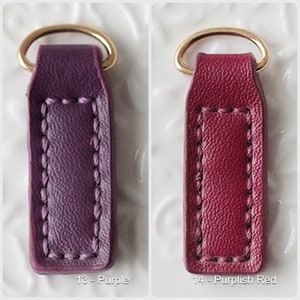 Op de afbeelding: Twee leren sleutelhangers, &eacute;&eacute;n paars en &eacute;&eacute;n purperrood, elk met een goudkleurige metalen ring. De sleutelhangers hebben een rechthoekige vorm met gestikte details en de nummers 13 en 14 met kleurnamen.