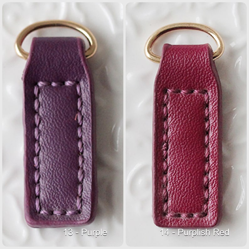 Op de afbeelding: Twee leren sleutelhangers, &eacute;&eacute;n paars en &eacute;&eacute;n purperrood, elk met een goudkleurige metalen ring. De sleutelhangers hebben een rechthoekige vorm met gestikte details en de nummers 13 en 14 met kleurnamen.