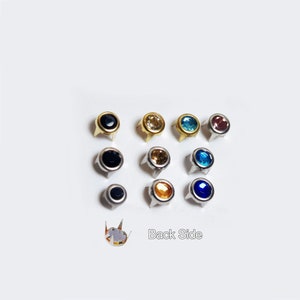 May include: Assortment of ten round, silver-toned, decorative nailheads with coloured glass rhinestones. The colours include black, gold, light blue, dark blue, and orange. The back side of one nailhead is visible, showing the prongs.