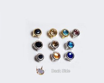 May include: Assortment of ten round, silver-toned, decorative nailheads with colored glass rhinestones. The colors include black, gold, light blue, dark blue, and orange. The back side of one nailhead is visible, showing the prongs.