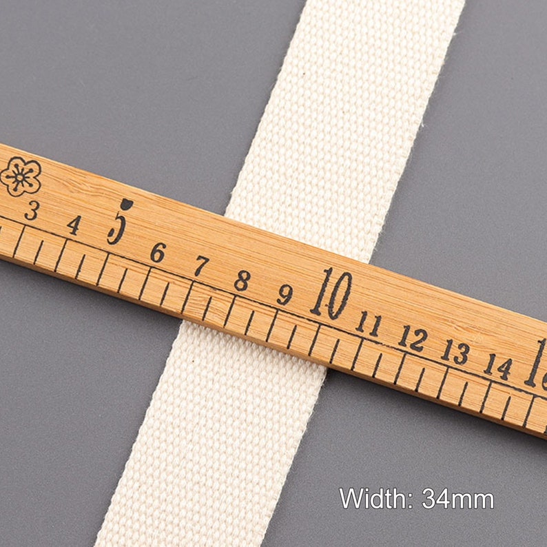 May include: A close-up of a white cotton webbing measuring 34mm wide. The webbing is lying on a wooden ruler with black markings.