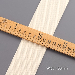 May include: A close-up of a natural white cotton webbing measuring 50mm wide. The webbing is laid on top of a wooden ruler with black markings.