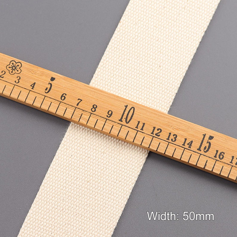 May include: A close-up of a natural white cotton webbing measuring 50mm wide. The webbing is laid on top of a wooden ruler with black markings.