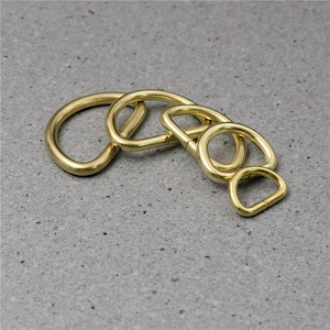 Puede incluir: Cuatro anillos en D de metal dorado de diferentes tama&ntilde;os. Los anillos est&aacute;n dispuestos en semic&iacute;rculo sobre una superficie gris texturizada.