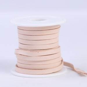 May include: A spool of light pink leather cord wound around a white plastic spool. The cord is about 1/4 inch wide.