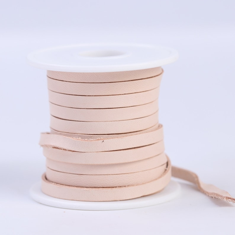 May include: A spool of light pink leather cord wound around a white plastic spool. The cord is about 1/4 inch wide.
