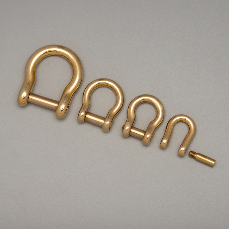 May include: Four gold-colored D-shackles of varying sizes are arranged on a gray surface. The shackles are U-shaped with a pin closure. One shackle is disassembled, showing the pin. The metal has a polished finish.