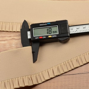 May include: A close-up of a beige elastic trim with a ruffled edge. A digital caliper measures the trim at 18 mm.
