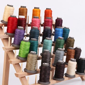 Könnte beinhalten: Ein Holzregal mit zahlreichen Garnrollen in verschiedenen Farben, darunter Rot, Orange, Grün, Blau und Braun. Die Spulen sind auf mehreren Ebenen angeordnet und präsentieren eine große Auswahl an Farbtönen für Näh- oder Bastelprojekte.