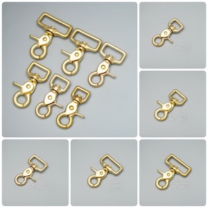 Puede incluir: Cinco mosquetones de metal dorado de diferentes tamaños. Los mosquetones están dispuestos en semicírculo sobre un fondo blanco.