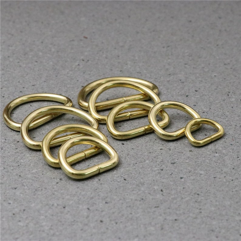 Puede incluir: Un conjunto de ocho anillos en D de metal dorado de diferentes tama&ntilde;os. Los anillos est&aacute;n dispuestos en una pila sobre una superficie gris texturizada.