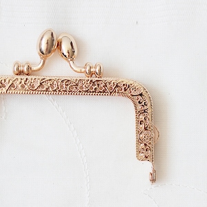 May include: Gold-toned metal purse frame with an ornate floral design. The frame has two rounded knobs at the top and a hook at the bottom.