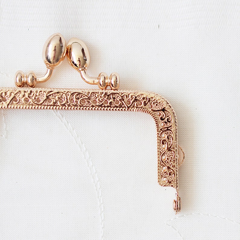 May include: Gold-toned metal purse frame with an ornate floral design. The frame has two rounded knobs at the top and a hook at the bottom.