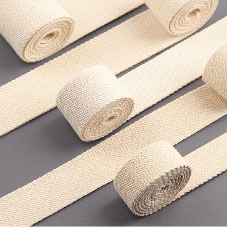 May include: Four rolls of natural beige cotton webbing, each rolled up and displayed on a gray surface. The webbing is woven with a subtle texture.