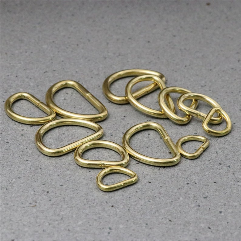 Puede incluir: Un conjunto de 10 anillos en D de metal dorado de varios tama&ntilde;os. Los anillos est&aacute;n dispuestos en un patr&oacute;n aleatorio sobre una superficie gris.