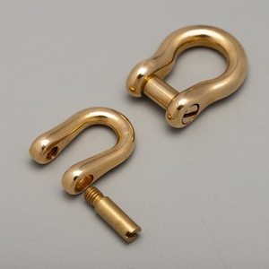 May include: Two gold-colored, U-shaped shackles, one assembled and one disassembled. The assembled shackle has a screw pin closure. The disassembled shackle shows the screw pin. The shackles are made of a shiny, metallic material.