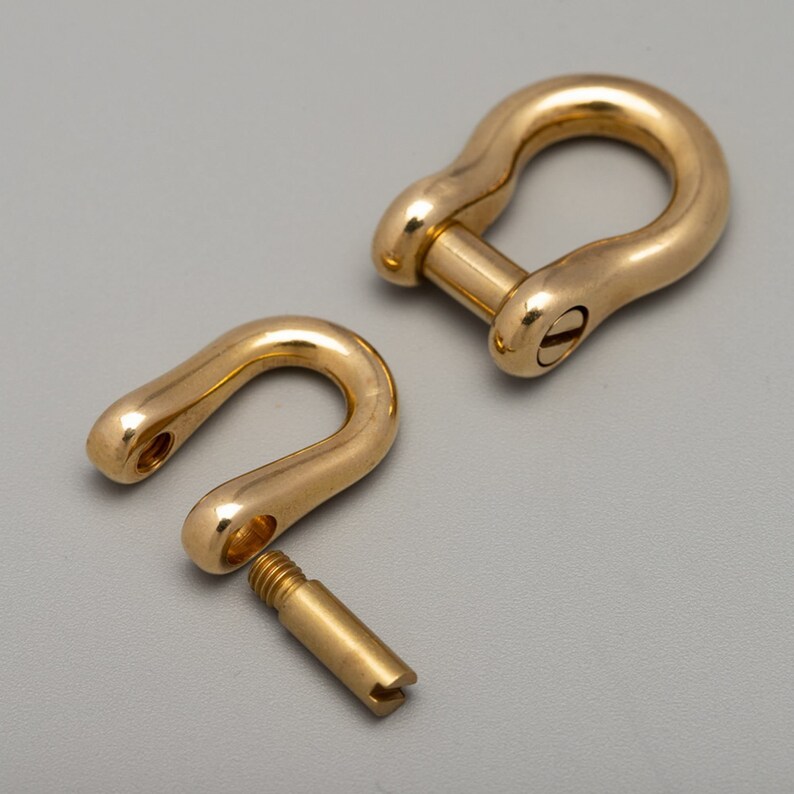 May include: Two gold-colored, U-shaped shackles, one assembled and one disassembled. The assembled shackle has a screw pin closure. The disassembled shackle shows the screw pin. The shackles are made of a shiny, metallic material.