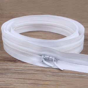 Puede incluir: Cinta de cremallera blanca con un tirador de cremallera blanco. La cinta de cremallera está enrollada y el tirador está en el centro del rollo.