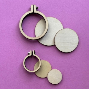 May include: Two wooden embroidery hoops with adjustable metal hardware and three wooden circles of varying sizes are displayed on a purple background. The hoops are light brown, and the circles are a natural wood colour.
