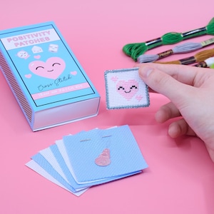 Op de afbeelding: Een kruissteekset met een blauwe doos met het opschrift "Positivity Patches". De doos heeft een lachend hart-ontwerp. Een afgewerkte patch met een hartontwerp wordt omhoog gehouden en er zijn verschillende blauwe stoffen vierkanten zichtbaar. Groen, grijs en bruin borduurgaren zijn ook aanwezig.