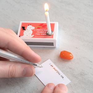 May include: A white matchbox with a red and white illustration of a dragon blowing fire. The matchbox has a small white candle with a flame on top. The text on the matchbox reads "A mighty flame follows a tiny spark" and "www.inamatchebox.com". A white card with the text "I am grateful for..." is on a white surface. A small orange stone is next to the card.