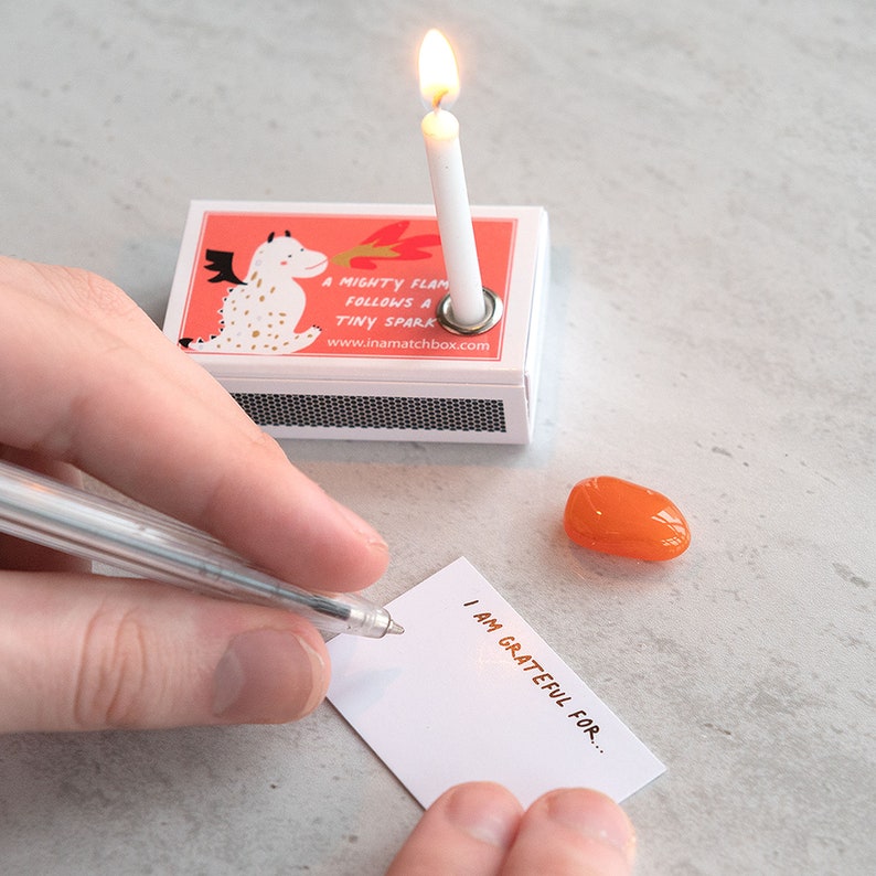 May include: A white matchbox with a red and white illustration of a dragon blowing fire. The matchbox has a small white candle with a flame on top. The text on the matchbox reads "A mighty flame follows a tiny spark" and "www.inamatchebox.com". A white card with the text "I am grateful for..." is on a white surface. A small orange stone is next to the card.